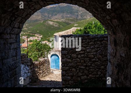 Ein malerischer Blick durch einen Steinbogen, der eine Kopfsteinpflasterstraße in der Altstadt von Himara enthüllt. Das Bild zeigt ein traditionelles Steinhaus mit Stockfoto