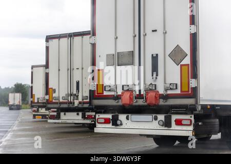 Reihe weißer Lkw mit Anhängertüren auf nassem Asphalt bei Regen unter bewölktem Himmel. Die Szene des Logistikzentrums zeigt den Gütertransport und die Sicherheitssiegel Stockfoto