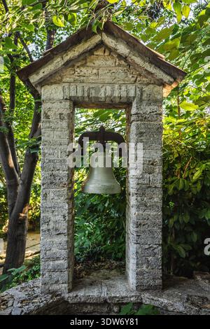 Alter Glockenturm aus Stein mit einer Metallglocke umgeben von üppigem Grün in einer ruhigen Gartenanlage. Stockfoto