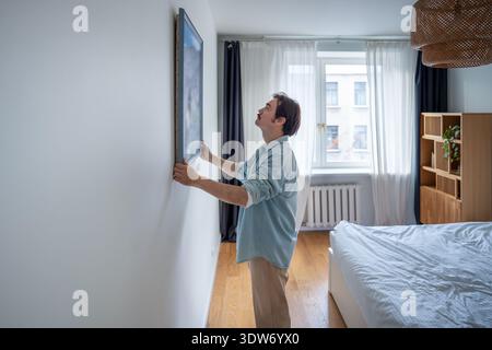 Fokussierte männliche Anordnung neuer Wohnkultur, hängende gerahmte Kunstwerke an weiße Wand in gemütlichem Schlafzimmer. Stockfoto