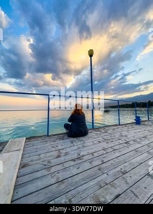 Issyk Kul See, Kirgisistan. Eine Frau sitzt auf einem hölzernen Pier und beobachtet den Sonnenuntergangshimmel, der sich in ruhigem Wasser spiegelt. Frau, die auf einem Pier sitzt und den Sonnenuntergang beobachtet Stockfoto