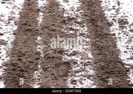 Parallel verlaufende Fahrzeugspuren bilden sich im schmelzenden Schnee wiederholende Linien, die die Erde darunter freilegen und die saisonalen Übergänge und die raue Winterstraße betonen Stockfoto