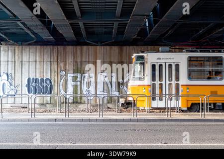 Gelbe Budapester Straßenbahn vorbei an der Graffiti-Mauer unter der Stadtüberführung, Ungarn. Stockfoto