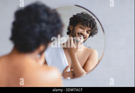 Junger Mann, der sich im Spiegel anfühlt und lächelt, während der Morgenroutine im Badezimmer Stockfoto