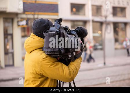 Kameramann mittleren Alters, der eine gelbe Jacke trägt und eine professionelle Videokamera auf einem Stativ in einer städtischen Straßenumgebung bedient. Stockfoto