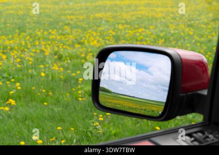 Seitenspiegel eines roten Autos, der eine wunderschöne Frühlingslandschaft mit blauem Himmel und Wolken über einem grünen Feld aus gelben Löwenzahn reflektiert. Stockfoto