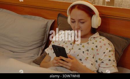 Junge Frau im Kopfhörer mit Smartphone im Bett Stockfoto
