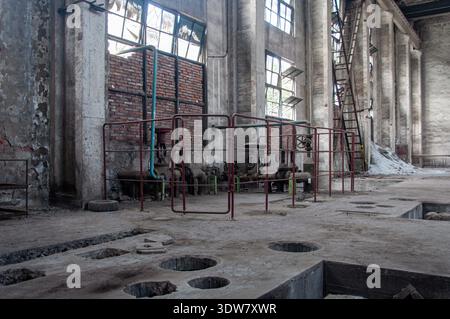 Horizontale Innenansicht einer verlassenen Fabrikhalle mit einem Betonboden mit Löchern, großen Fenstern, Rohren und verstreuten Trümmern, die einen Sinn erwecken Stockfoto