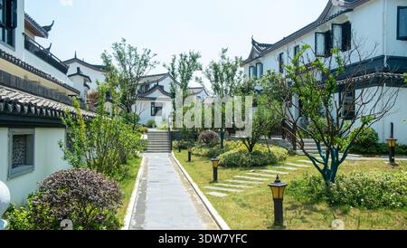 Horizontaler Blick auf einen gepflasterten Steinweg, der sich durch einen Landschaftsgarten in einer Gemeinschaft von weißen Wänden traditioneller chinesischer Häuser schlängelt. Stockfoto