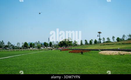 Weite Landschaftsaufnahme eines gepflegten grünen Rasenparks mit roten Blumenbeeten vor einem riesigen klaren blauen Himmel mit einer weit entfernten Drohne. Stockfoto
