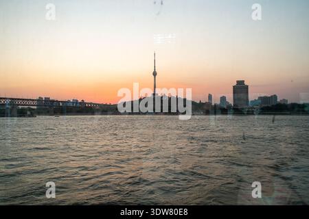 Breiter horizontaler Blick auf einen ruhigen Fluss bei Sonnenuntergang, mit einer Skyline der Stadt und einem markanten Fernsehturm vor dem warmen, glühenden Himmel. Stockfoto