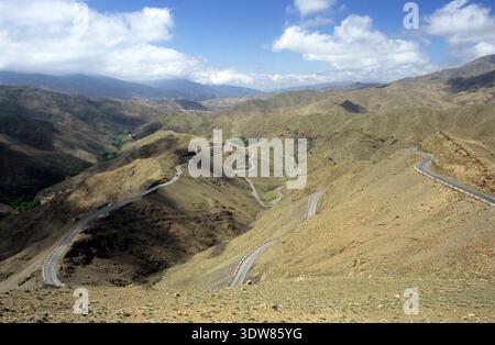 Die gewundene Bergstraße schlängelt sich durch die rauen, trockenen Hänge des Atlasgebirges unter einem verstreuten blauen Himmel, Marokko. Stockfoto