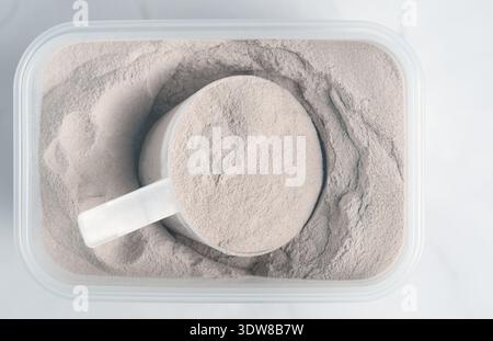 Überkopf-Schuss der Textur von Vanille- oder Molkenproteinpulver in einem Kunststoffbehälter mit Messlöffel Stockfoto