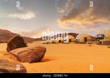 Kuppelzelte liegen entlang des Sandes vor der Kulisse von felsigen Hügeln. Die Sonne geht unter, wirft warmes Licht über die trockene Landschaft und schafft Schatten und Formen Stockfoto