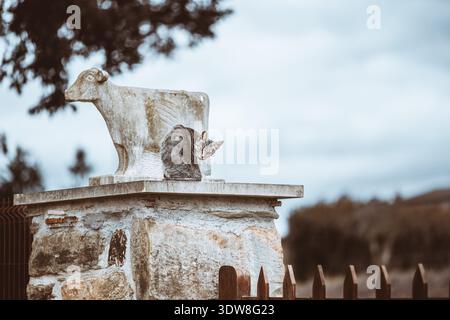 Neugierige Tabbykatze, die auf einer rustikalen Steintor-Säule neben einer verwitterten Kuh-Statue thront, auf die Kamera zurückblickend, ländliche Bauernstimmung, geringe Tiefe des Feldes, also Stockfoto