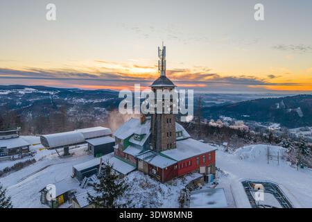 Der Aussichtsturm steht im Winter auf dem Tanvaldsky Spicak Berg. Schnee bedeckt die Strukturen, während der Sonnenuntergang den Himmel erleuchtet. Skier und Snowboards sind in der Gegend zu sehen. Stockfoto