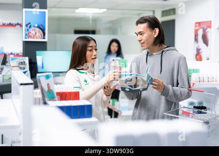 Asiatischer Apotheker erklärt verschiedene Produkte auf Flyer mit männlichen Kunden in der modernen Apotheke. Freundliches medizinisches Fachpersonal bietet aufmerksame Beratung und baut Vertrauen während der medizinischen Beratung auf. Stockfoto