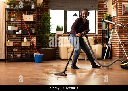 Der fröhliche Hausbesitzer trägt Kopfhörer, während er das Apartment mit Musik und Staubsauger reinigt und sich um die Wochenendarbeiten kümmert. Glückliche afroamerikanerin saugt das Wohnzimmer und räumt das Haus auf. Stockfoto