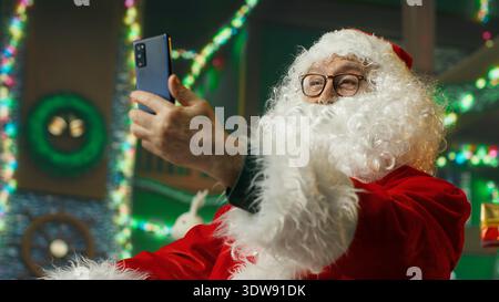 Fröhlicher Weihnachtsmann, der mit Kindern über das Smartphone Videoaufnahmen macht, bringt Freude. Weihnachtsmann unterhält Kinder während der Telefonkonferenz, verbreitet das Gefühl des Staunens während Weihnachten, Kamera A Stockfoto