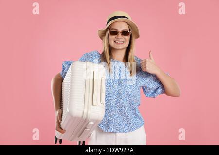 Glückliche junge Frau mit Koffer, die Daumen hoch auf rosa Hintergrund zeigt Stockfoto
