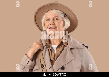Ältere Frau in stilvollem Trenchcoat mit Hut auf beigefarbenem Hintergrund Stockfoto