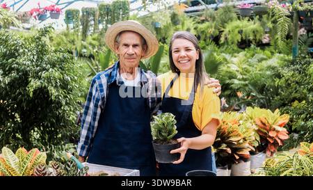 Familienbetrieb im Frühjahrsgarten im Gewächshaus - hispanische Seniorenbauerin und lateinamerikanische Frau mit Topfpflanze Stockfoto