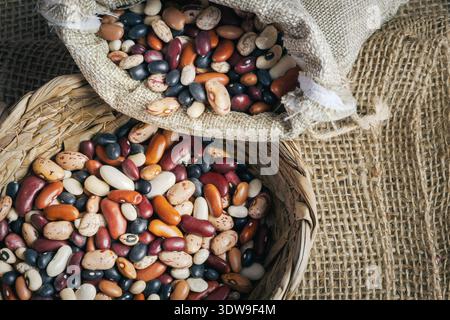 Ein kleiner, runder, gewebter Korb und ein Sacksack sind gefüllt mit einer bunten Mischung aus verschiedenen ungekochten getrockneten Bohnen und Hülsenfrüchten, i Stockfoto