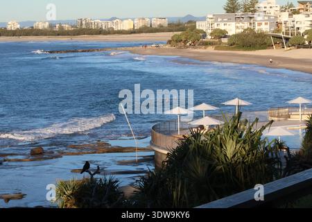 Surfer auf einer Welle in der Nähe eines Aussichtspunkts an der Küste mit Sitzplätzen im Freien und Sonnenschirmen mit Blick auf das Meer. Stockfoto