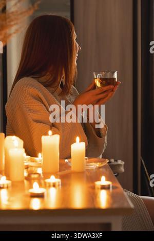 Frau im Bademantel genießt heißen Tee, beleuchtet durch flackernde Kerzen, findet Ruhe und Ruhe. Wellness und Spa-Abend Stockfoto