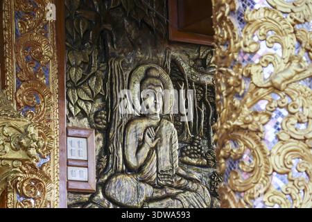 Heitere spirituelle Holzskulptur mit Buddha-Schnitzerei an der Tempelwand in Asien. Komplexe Gold- und braune Kunstwerke zeigen traditionelles Kunsthandwerk und Ruhe Stockfoto