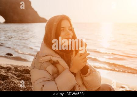 Frau, Apfel, Strand. Eine junge Frau in einer geschwollenen Jacke hält einen Apfel, während sie den wunderschönen Sonnenuntergang über dem ruhigen Meer beobachtet. Stockfoto
