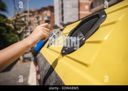 Werfen Sie an einem sonnigen Tag in der Stadt eine Plastikflasche von Hand in einen Recyclingbehälter mit gelbem Deckel. Stockfoto