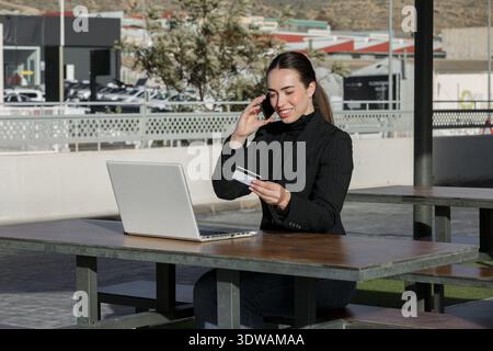 Eine Frau sitzt an einem Tisch im Freien, benutzt einen Laptop und spricht am Telefon. Sie hält eine Kreditkarte in der rechten Hand. Diese Szene spielt in einem Stockfoto