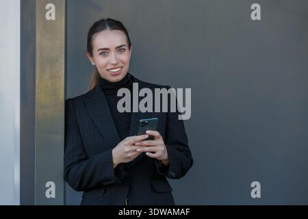 Eine Frau mit schwarzem Blazer lehnt sich an eine graue Wand, während sie ihr Smartphone benutzt. Sie scheint glücklich und engagiert zu sein. Die Umgebung lässt einen modernen urba vermuten Stockfoto