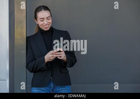 Eine Frau steht vor einer grauen Wand, während sie mit ihrem Smartphone beschäftigt ist. Sie trägt einen Nadelstreifenanzug und lächelt, während sie mit dem Gerät interagiert, wahrscheinlich c Stockfoto