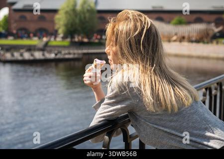 Entspannte positive Frau, die am Sommertag Eiscreme isst und über den Fluss von der Brücke aus beobachtet. Stockfoto