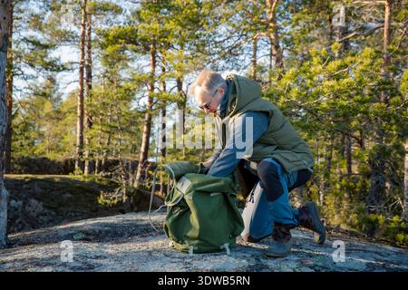 Reifer Mann in seinen 50ern, der zur goldenen Stunde einen Vintage-Rucksack im nordischen Wald überprüft. Aktiver Outdoor-Lifestyle im Midlife Stockfoto