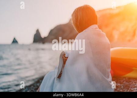 Person, Strand, Sonnenuntergang. Eine Person, die in ein Handtuch gewickelt ist, sitzt friedlich an einem felsigen Strand und genießt den ruhigen Sonnenuntergang am Meer. Stockfoto