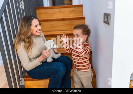 Mutter und Kleinkind lächeln und spielen mit Teddybären auf der Treppe zu Hause Stockfoto