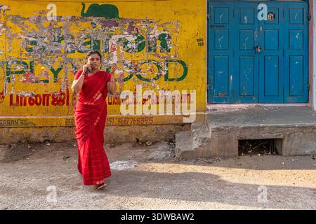 Indische Frau mit traditionellem rotem Saree mit Gesichtspiercings, die neben einer hellgelben Wand und einer blauen Holztür in Indien steht. Stockfoto