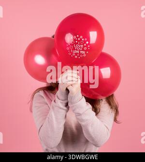 Valentinstag-Konzept. Frau, die ihr Gesicht mit drei roten Ballons bedeckt, mit den Worten I Love You isoliert auf rosa Hintergrund || Model veröffentlicht Stockfoto