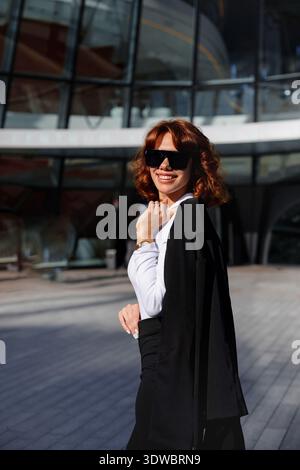 Glückliche Geschäftsfrau, die im Sonnenlicht lächelt, während sie in der Nähe eines modernen Bürogebäudes mit entspannter, selbstbewusster Pose steht. Stockfoto