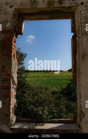 Blick durch verlassene Backsteinfenster in Richtung grasbewachsener Küstenfelder unter klarem Himmel auf Rhodos, Griechenland. Stockfoto