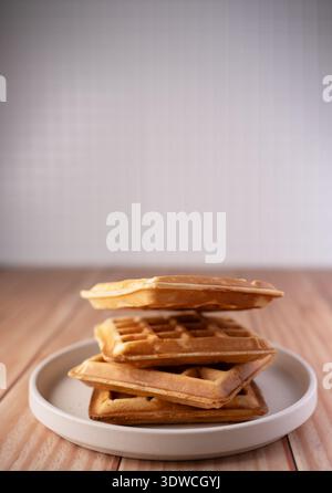Stapel goldener Waffeln auf einer weißen Keramikplatte auf einer Holzplatte in minimaler Küchenatmosphäre, warmen neutralen Tönen und sanftem natürlichem Licht mit soll Stockfoto