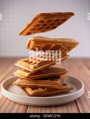 Goldbraune Waffeln, die über einer weißen Keramikplatte schweben, auf einer Holzplatte mit neutraler Fliesenunterseite, warmer sanfter Beleuchtung, Stockfoto