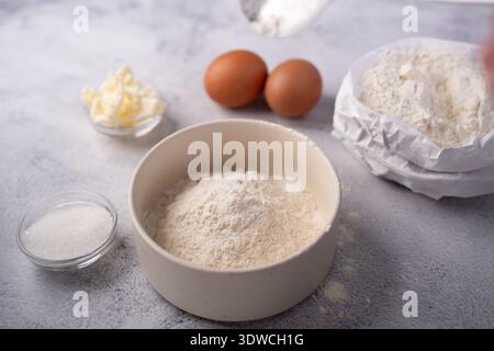 Nahaufnahme der Backzutaten auf einer strukturierten Arbeitsplatte: Eine beige Schüssel Mehl, Papiertüte Mehl, zwei braune Eier, kleine Schüsseln Zucker und Butter, Stockfoto