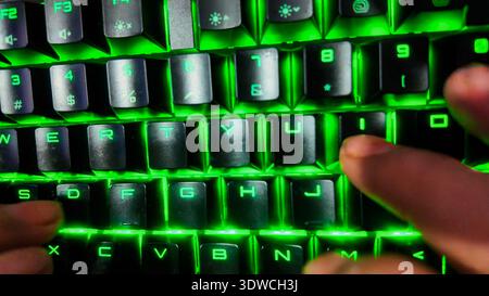 Eine Nahaufnahme einer Person aus der Nähe, die auf einer schwarzen mechanischen Tastatur mit dynamischer RGB-Hintergrundbeleuchtung in einem schwach beleuchteten Raum tippt. Im Mittelpunkt steht Stockfoto