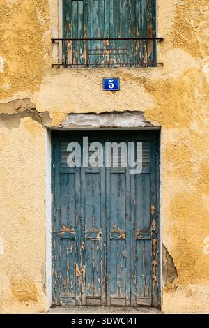 Rustikale, türkisfarbene Fensterläden und Tür an einer verwitterten ockerfarbenen Gebäudefassade, mit einer blauen Hausnummerntafel mit der Aufschrift '5', die an rustikalen französischen Charme erinnert Stockfoto