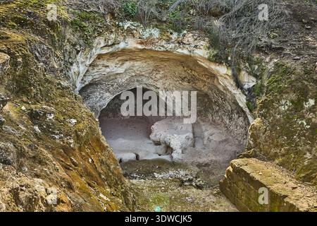 Natürlicher Höhleneingang mit felsigen Mauern und bewachsener Vegetation, der in ein dunkles Inneres führt, umgeben von erdbem Gelände und Steintreppen in einem ländlichen Raum Stockfoto