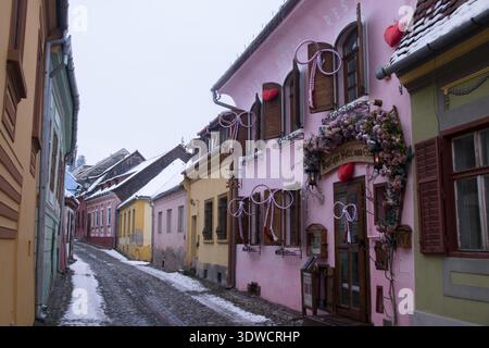 Die Menschen spazieren durch die engen Gassen von Sighisoara und sehen farbenfrohe Häuser und eine leichte Schneedecke im Winter in Siebenbürgen. Stockfoto